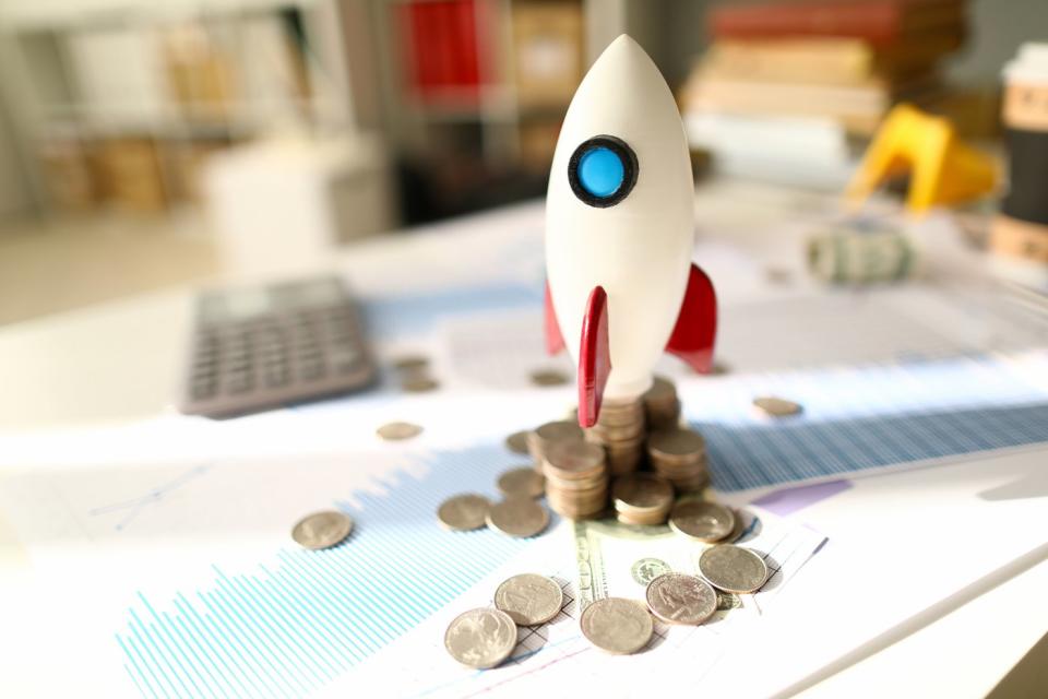 A toy rocket sits on top of messy piles of coins and documents displaying financial data. 